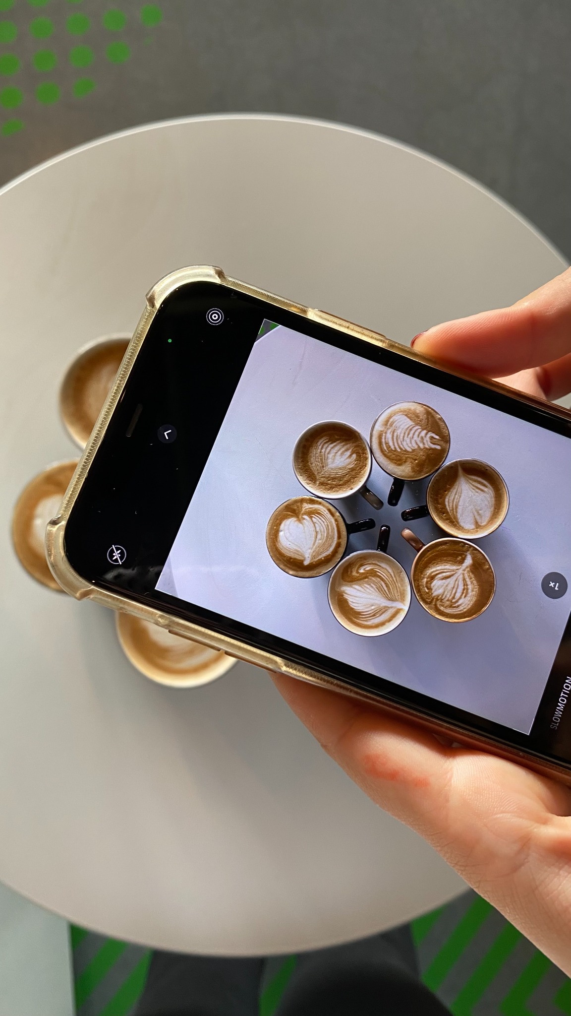 A hand holds a phone photographing latte cups on a round table.